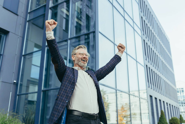 Successful and happy gray-haired boss outside office building rejoices success and victory, triumphs with hands up, businessman business owner in business suit and glasses, mature investor banker. - Photo, Image