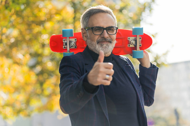 Positive caucasian bearded gray-haired guy in his 50s showing thumbs up to the camera while holding bright-orange mini skateboard behind his neck. Autumn park. Outdoor shot. Blurred yellow tree leaves - Фото, изображение