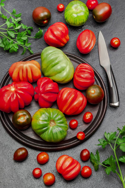 Red and green tomatoes on cutting board and on table. Parsley sprigs and knife. Flat lay. Black background. - Foto, Bild