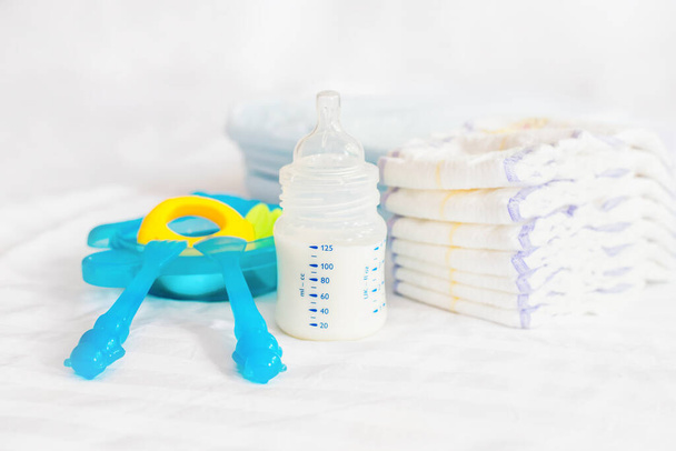 赤ん坊のプラスチック皿のセット。ミルクボトル、ニンブラー、ブルーカトラリー、スプーン、フォーク、オムツ、おもちゃ、使い捨ておむつを白いベッドの背景に供給しています。健康的な栄養。正面図 - 写真・画像