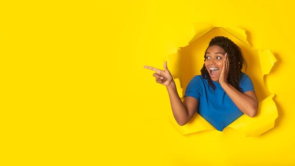 Emocionada mujer afroamericana señalando dedo aparte de espacio vacío publicidad Wow oferta posando en el agujero en el papel amarillo roto, fondo del estudio. Mira allí Concepto. Panorama - Foto, imagen