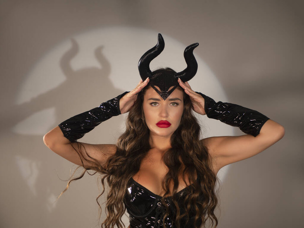 Young brunette with red lips and long wavy hair adjusting devil horns and looking at camera against gray background - Fotoğraf, Görsel