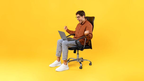Joyful Guy Shaking Fists Using Laptop Computer Celebrating Business Success Sitting In Office Chair Working Online In Studio Over Yellow Background. Panorama, Side View Shot - Photo, image