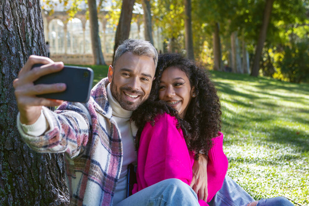 Gülümseyen ve mutlu çift parkta çimlerin üzerinde oturmuş cep telefonlarıyla fotoğraf çekiyorlar.  - Fotoğraf, Görsel
