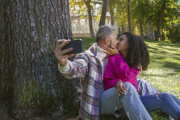 happy couple sitting on the grass in the park kissing and taking a picture with the phone, - 写真・画像