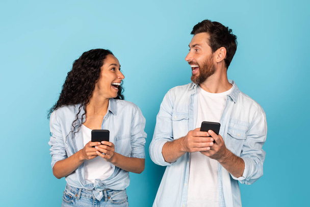 Překvapen šťastný vzrušený mladý různorodý pár s chytrými telefony se na sebe podívají, radují se z online vítězství, izolované na modrém pozadí, studio. Lidé emoce, dobré zprávy, skvělá nabídka, reklama a prodej - Fotografie, Obrázek