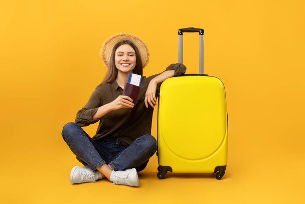 Vacaciones. Mujer feliz Turista sentada cerca de la maleta sosteniendo pasaportes y boletos de viaje posando con sombrero en el fondo del estudio amarillo. Concepto de turismo - Foto, Imagen