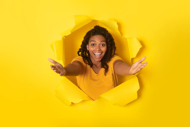 Izgalmas afro-amerikai nő nyújtózkodó karok lyukon keresztül szakadt sárga papír pózol mosolygós kamera a stúdióban. Wow Ajánlat, Kereskedelmi Reklám koncepció - Fotó, kép