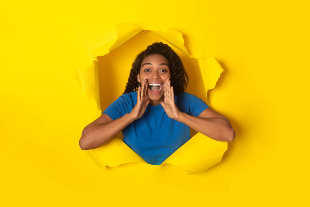 Emocionální Afroameričanka křičí při pohledu skrz díru v roztrhaném žlutém papíru reklama Skvělá nabídka při pohledu na kameru Posing On Yellow Studio pozadí. Nápis - komerční reklama - Fotografie, Obrázek