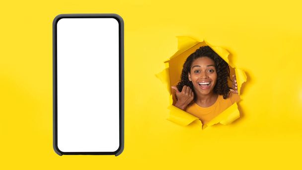 Emocionada mujer afroamericana señalando el teléfono inteligente con enorme pantalla vacía publicidad Wow oferta móvil posando en papel amarillo roto, señalando dedo en el teléfono inteligente grande. Burla. - Foto, imagen