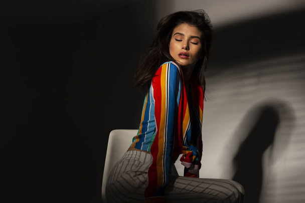 Portrait of a young seductive brunette woman in colorful top. Image with shadows, posing with closed eyes. - Foto, Imagen