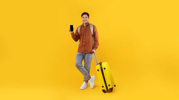 App de viagem. Turista masculino mostrando tela em branco do telefone recomendando oferta móvel para viajantes sorrindo para câmera posando de pé com mala sobre fundo de estúdio amarelo. Panorama - Foto, Imagem