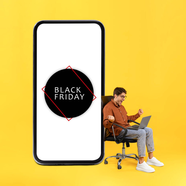 Black Friday Verkaufsangebot. Emotionaler Typ mit Laptop, der Ja gestikuliert und schreit, wenn er in der Nähe eines riesigen Handys mit Rabattwerbung auf gelbem Hintergrund im Studio sitzt. Vierecksschuss - Foto, Bild