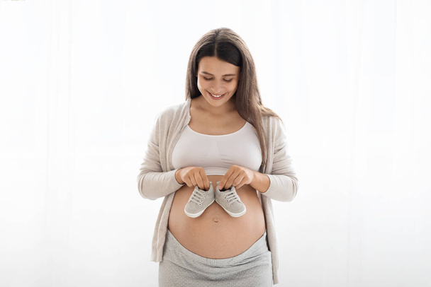 Fröhliche junge schwangere Frau in bequemer Homewear steht vor weißem Hintergrund, hält niedliche Babyschuhe auf ihrem großen Bauch und lächelt, werdende Mutter wartet auf ihr Kind, Kopierraum - Foto, Bild