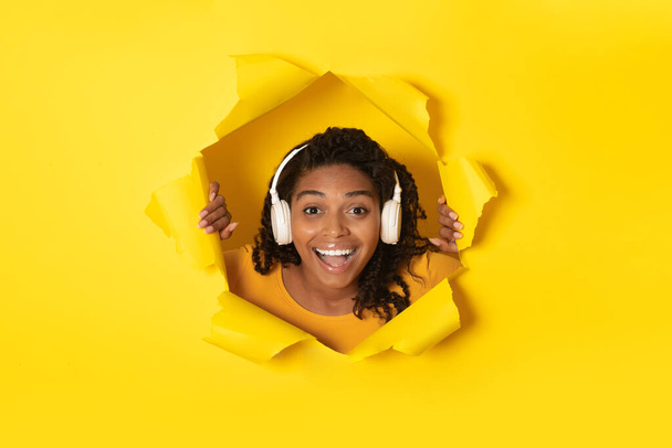 Радісна чорна леді носить бездротові навушники, що слухають музику, посміхаючись, дивлячись через отвір у жовтому папері, рекламному музичному додатку. Жінка насолоджується улюбленою піснею
 - Фото, зображення
