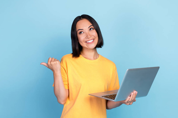 Portrait of nice good mood interested girl dressed yellow t-shirt directing empty space arm hold laptop isolated on blue color background. - Foto, immagini