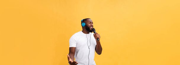 Portret wesołego pozytywnego szyku. przystojny Afrykanin człowiek trzyma mikrofon i mając słuchawki na głowie słuchanie muzyki śpiew piosenka ciesząc weekend wakacje odizolowany na żółtym tle. - Zdjęcie, obraz