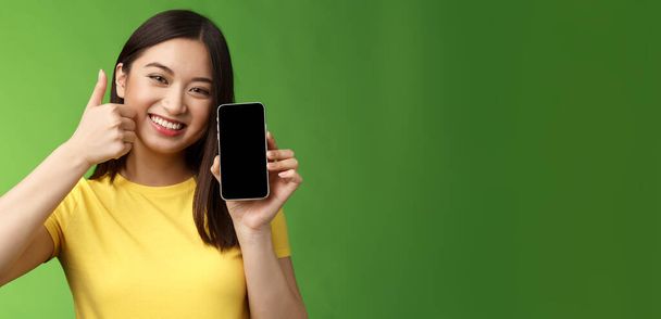 クローズアップ満足かわいいアジアの女の子は肯定的なフィードバックを与えます,スマートフォンの画面を示す親指アップ,素晴らしい新しいアプリを紹介します,良いアプリケーションをお勧めします,ゲームでの自慢の暴行スコア. - 写真・画像