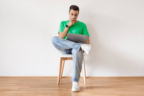 Pensive arab άνθρωπος χρησιμοποιώντας φορητό υπολογιστή και τη σκέψη, περιήγηση στο διαδίκτυο, ενώ κάθεται στην καρέκλα κατά λευκό τοίχο στο σπίτι. Ανεξάρτητος επαγγελματίας στη Μέση Ανατολή που εργάζεται online στον υπολογιστή - Φωτογραφία, εικόνα
