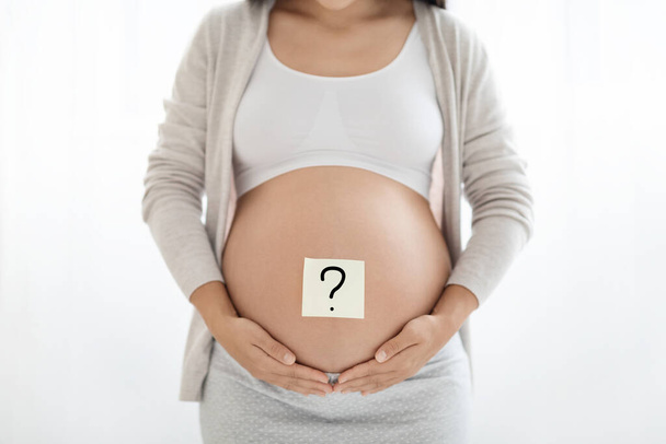 Conceito de escolher o nome do bebê. Mulher grávida irreconhecível em roupa de casa com ponto de interrogação no papel adesivo em sua barriga grande, abraçando sua barriga grande, fundo branco - Foto, Imagem