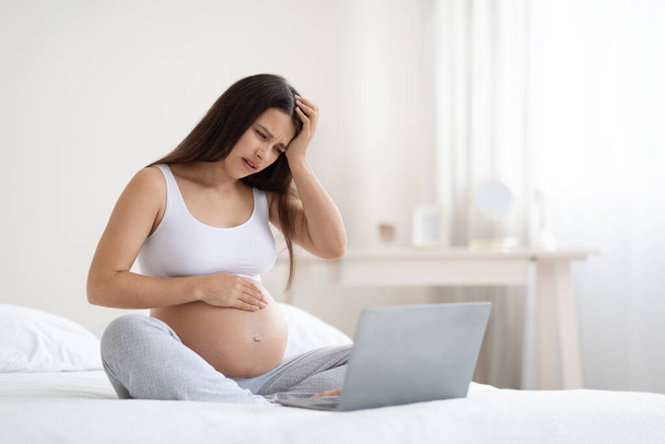 Nemocná mladá těhotná žena sedí na posteli před notebookem, dotýká se svého velkého břicha a hlavy, trpí migrénou, má videohovor s lékařem při pobytu doma, panorama s kopírovacím prostorem - Fotografie, Obrázek