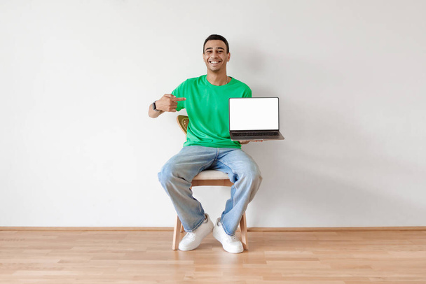 Király honlap. Pozitív arab fickó laptopra mutat fehér üres képernyővel, széken ül a fehér stúdió falán, mockup. Vidám srác hordozható számítógéppel - Fotó, kép