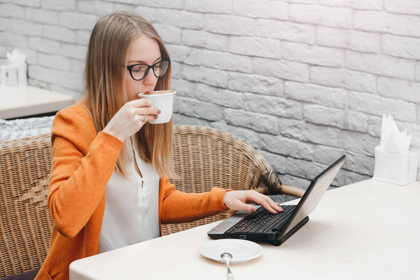 Blondýnka v kavárně s laptopem a šálkem kávy. Mladá dívka na volné noze pracuje na notebooku - Fotografie, Obrázek