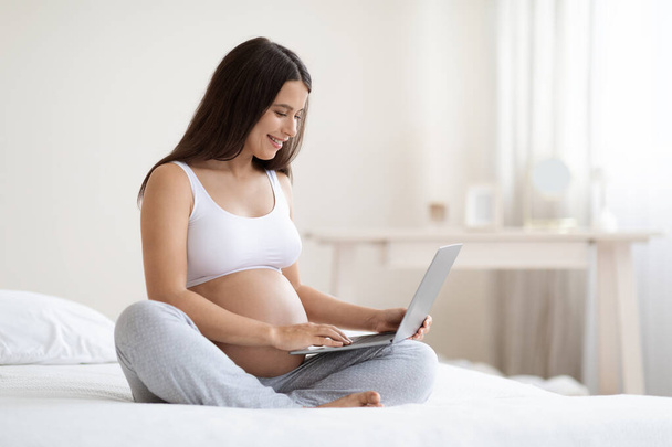 Radostné atraktivní očekává žena v pyžamu relaxaci v posteli s notebookem doma, dlouhosrstá těhotná žena surfování na internetu, čtení blog pro těhotné ženy, ložnice interiér, kopírovací prostor - Fotografie, Obrázek