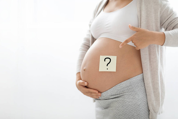 Ongeboren baby geslacht concept. Gehakt shot van zwangere vrouw in homewear gericht op sticker met vraagteken op haar buik, witte studio achtergrond, panorama met kopieerruimte - Foto, afbeelding