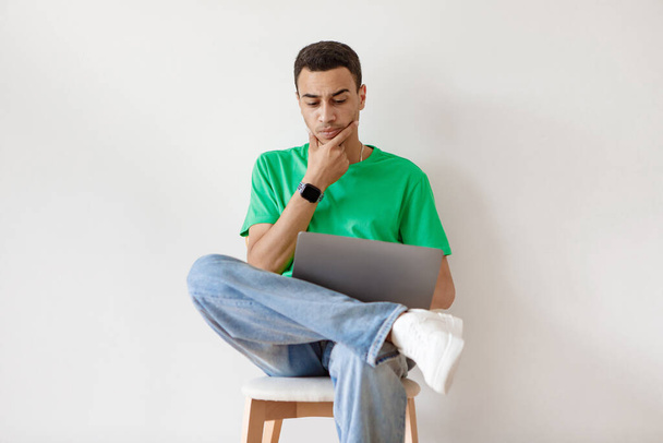 Bedachtzame arabische man die laptop gebruikt terwijl hij op een stoel tegen een witte muur zit, werkt of studeert op afstand op een draagbare pc. Pensive man communiceert online - Foto, afbeelding