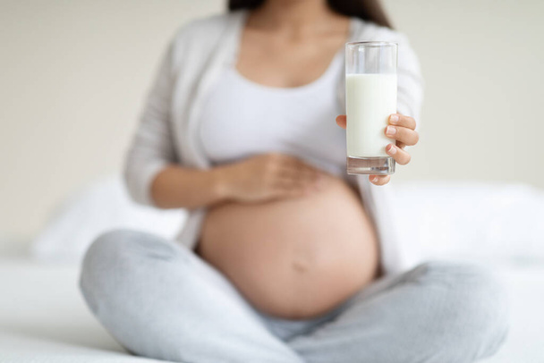 Copo de leite fresco na mão da mulher grávida, irreconhecível esperando senhora em casa sentada na cama em casa, tocando sua barriga grande, bebendo leite. Nutrição durante o conceito de gravidez - Foto, Imagem