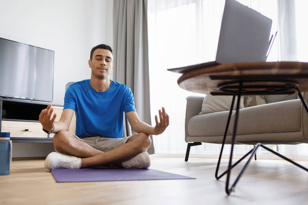 Domestic yoga. Arab man meditating in front of laptop at home, sitting with closed eyes in lotus position, enjoying online domestic trainings - Photo, Image