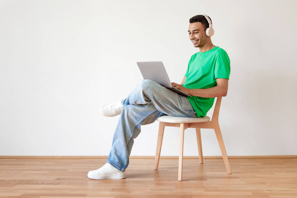 Jonge Arabische man die pc gebruikt, online op laptop werkt en een draadloze hoofdtelefoon draagt, op een stoel over een witte muur zit, kopieert ruimte. Guy kijken film film of chatten online - Foto, afbeelding