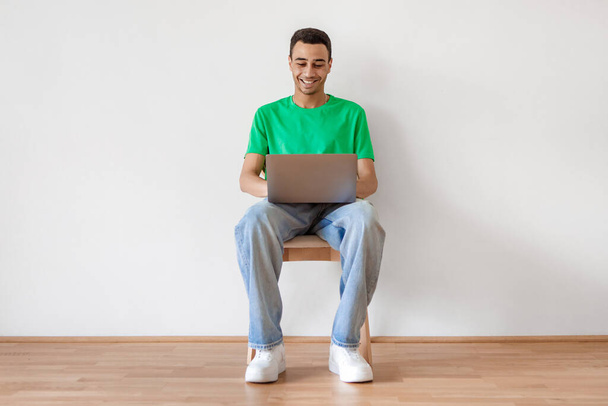 Cheerful arab male freelancer using laptop computer, sitting on chair over white wall and typing on keyboard, copy space. Remote job concept - Photo, Image