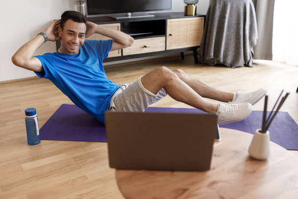 Sport and healthy lifestyle. Young arab man holding hands behind his head and doing exercises for abdominal muscles, watching online training on laptop, exercising at home - Photo, Image