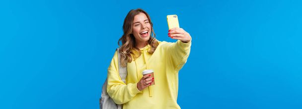 Lifestyle, university and education concept. Cheerful good-looking blond female student or traveller taking selfie with backpack and take-away coffee in paper cup, wink to mobile phone camera. - 写真・画像