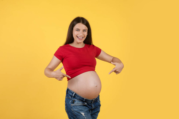 Vrolijke Zwangere vrouwelijke wijzende vingers op haar buik en glimlachen bij de camera terwijl poseren geïsoleerd op gele achtergrond, Happy Young Expectant vrouw aangeeft haar grote buik, Genieten van zwangerschapstijd - Foto, afbeelding