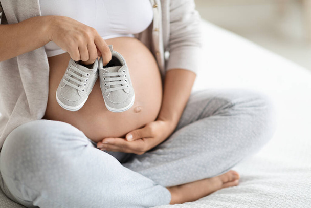 Обрезанная беременной женщиной в удобной домашней одежде, сидящей дома на кровати, держа милые детские туфельки на своем большом животике, неузнаваемая будущая мать, ждущая ребенка, копирующее пространство - Фото, изображение