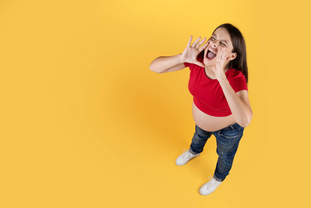Над видом молодая беременная женщина с руками у рта сделать объявление, ожидать женского обмена новостями или информацией, находясь в изоляции на фоне желтой студии, скопировать пространство - Фото, изображение