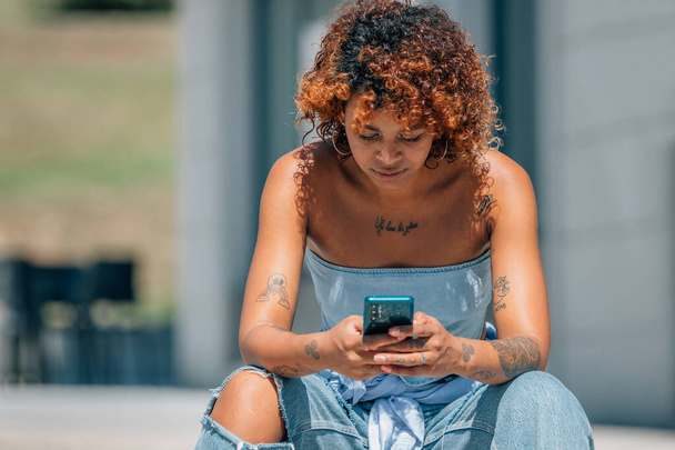 miejski latynoski afrykański dziewczyna amerykański patrząc na telefon komórkowy siedzi na ulicy na zewnątrz - Zdjęcie, obraz