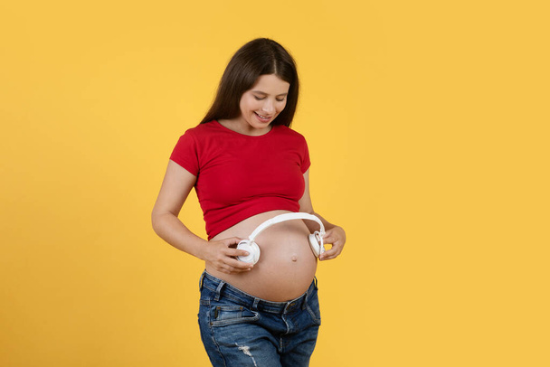 Mujer embarazada joven feliz sosteniendo auriculares inalámbricos cerca del vientre, sonriendo hembra embarazada en ropa casual tocando música para el bebé en el vientre mientras está de pie aislado en el fondo amarillo, espacio de copia - Foto, Imagen