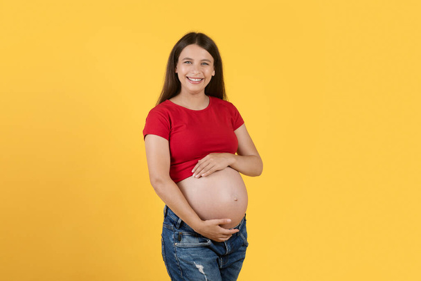 Piękna młoda kobieta Obejmuje jej duży brzuch w ciąży i uśmiecha się do aparatu, Happy Expectant Kobieta czułe dotykanie brzucha, stojąc odizolowany na żółtym tle Studio, Kopiuj przestrzeń - Zdjęcie, obraz