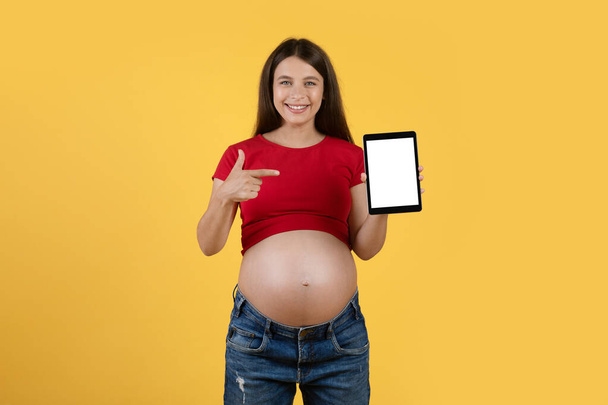 Smiling Pregnant Woman Showing Digital Tablet With Blank Screen And Pointing On It, Cheerful Expectant Female Demonstrating Free Copy Space For App Or Website Design, Yellow Background, Mockup - Фото, изображение