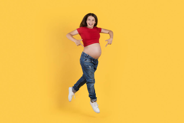 Radosna młoda kobieta w ciąży wskazując na jej brzuch podczas skakania w powietrzu nad żółtym tle w studio, pozytywne aktywne kobiety korzystające z aktywnego stylu życia i zdrowej ciąży, przestrzeń skopiować - Zdjęcie, obraz