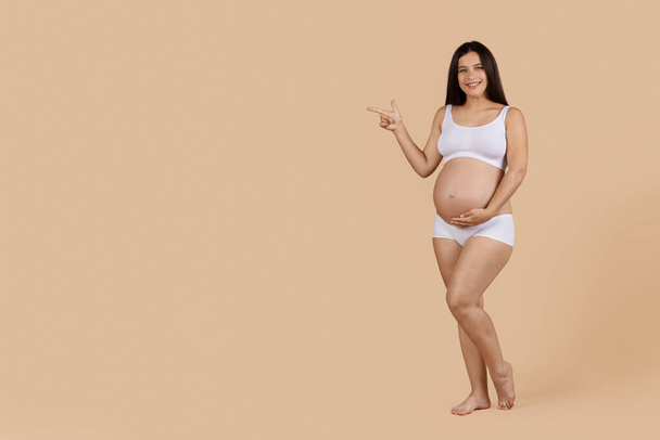 Schwangerschaftsanzeige. Schöne lächelnde Schwangere in Unterwäsche, die auf Kopierplatz zeigt, während sie über beigem Studiohintergrund posiert, glückliche werdende Frau, die freien Platz für Werbung zeigt - Foto, Bild