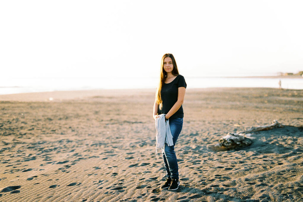 Chica en jeans con un suéter en sus manos se encuentra en una playa de arena junto al mar. Foto de alta calidad - Foto, imagen