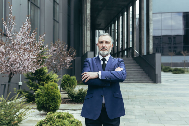 Portrait of senior handsome man, a businessman, a judge, a lawyer. Standing in a suit near the courthouse, office center. Crossed his arms in front, looks at the camera, serious. - Fotografie, Obrázek