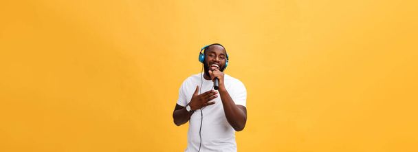 Retrato de alegre chic positivo. hombre africano guapo sosteniendo micrófono y teniendo auriculares en la cabeza escuchando música cantando canción disfrutando de vacaciones de fin de semana aislado sobre fondo amarillo. - Foto, Imagen