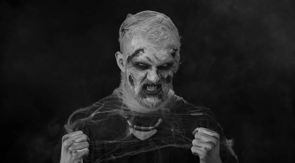 Homme effrayant avec Halloween zombie ensanglanté maquillage blessé, essayant d'effrayer, expressions du visage. Des cris. Sinistre mort-vivant isolé sur fond noir studio. Des rituels vaudous. Art corporel mode - Photo, image