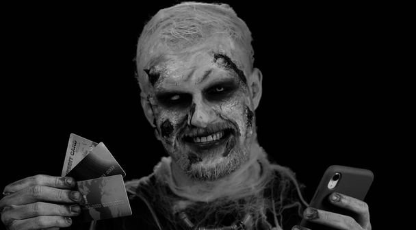 Sinister man with horrible scary Halloween zombie make-up using credit bank cards and mobile phone, transferring money purchases online shopping. Dead guy with wounded bloody scars face, black room - Photo, Image